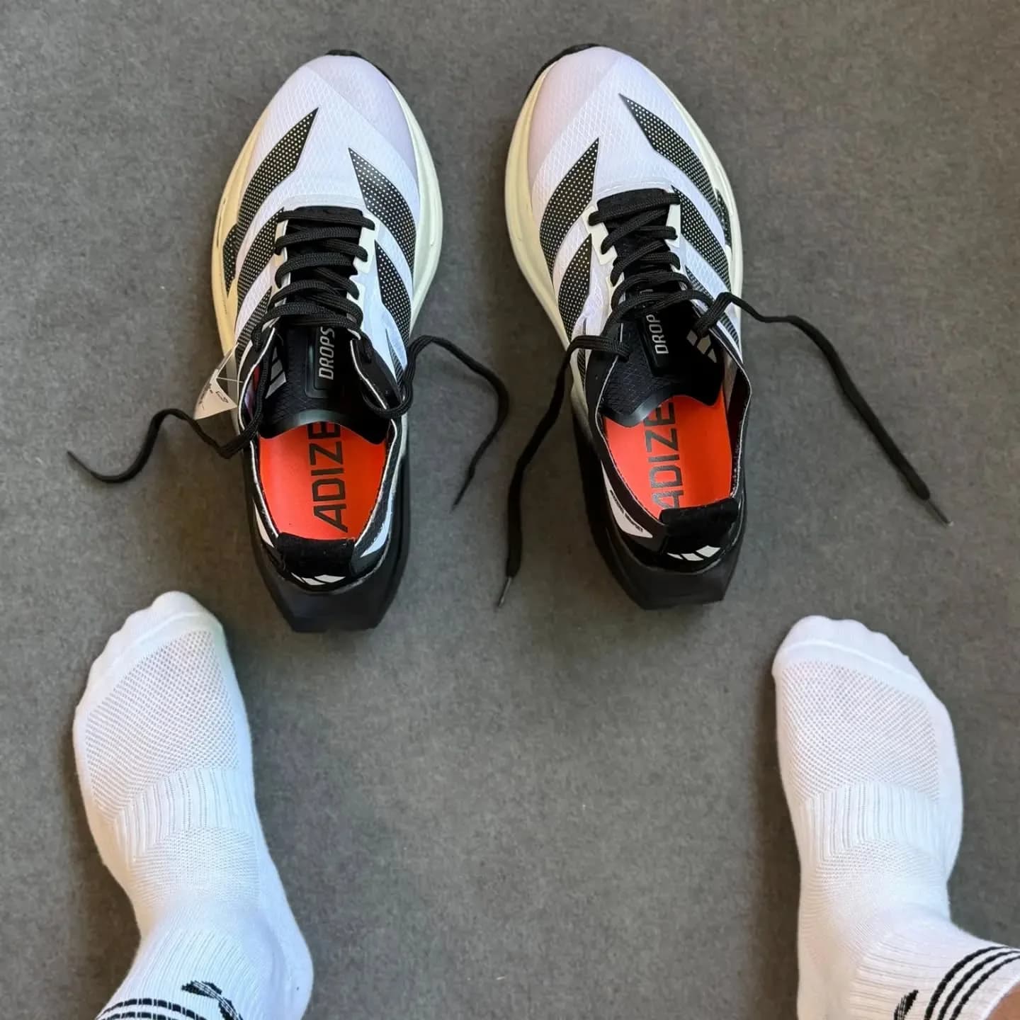 Um par de Adidas Adizero, branco com a sola vermelha, visto de cima entre dois pés de meia branca